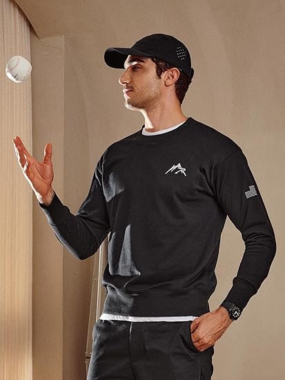 A male model wearing a black crewneck sweatshirt with a mountain logo and flag sleeve detail, casually tossing a baseball in the air.